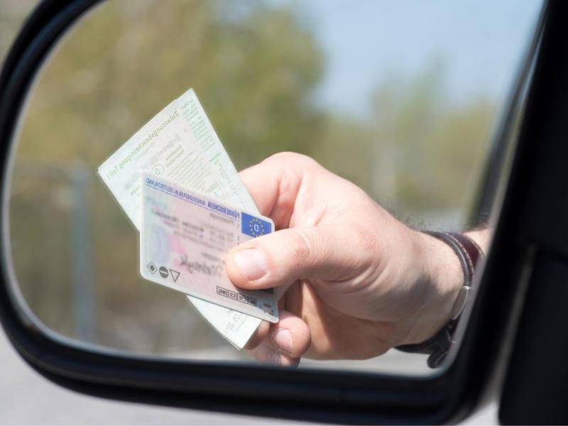 Person hält einen Führerschein und ein Ausweisdokument aus dem Auto, die im Seitenspiegel zu sehen sind.