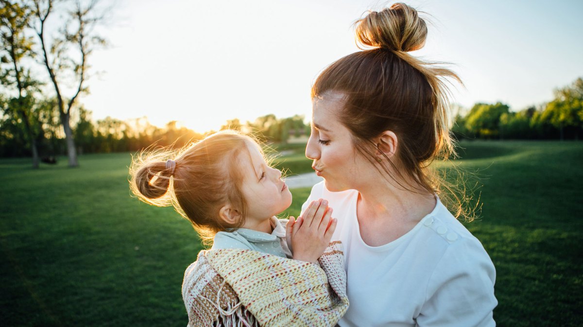 Mutter und Tochter (4) draußen im Park. Die Mutter trägt die Tochter, die in eine Decke gewickelt ist.