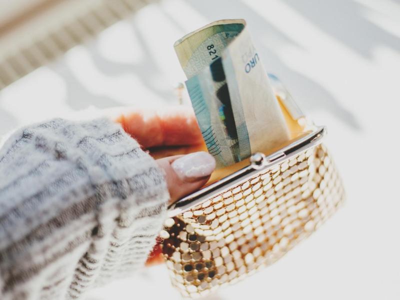 Frau mit künstlichen Gelnägeln hält eine goldene Geldbörse, aus der Geldscheine gucken.