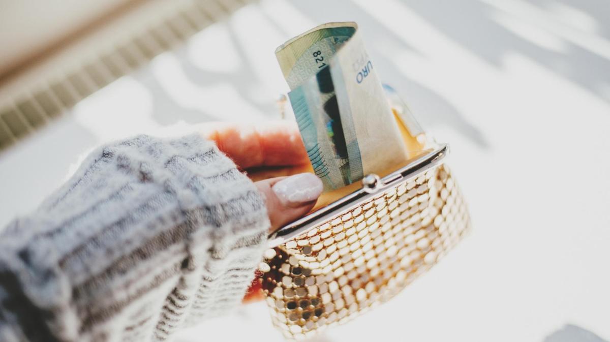 Frau mit künstlichen Gelnägeln hält eine goldene Geldbörse, aus der Geldscheine gucken.