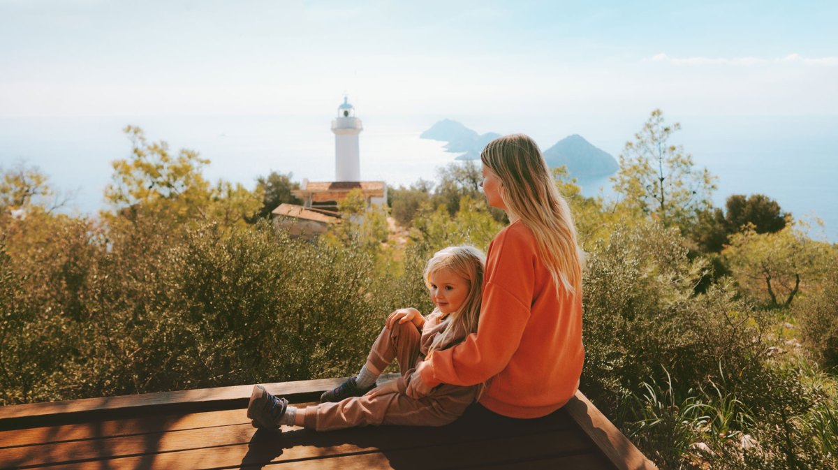 Mutter und Kind auf Reisen, sitzen auf einem Felsvorsprung, im Hintergrund erkennt man einen Turm und Berge im Meer.