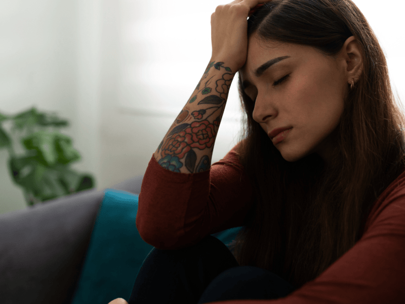 Frau mit Tattoos am Arm sitzt traurig auf einem Sofa