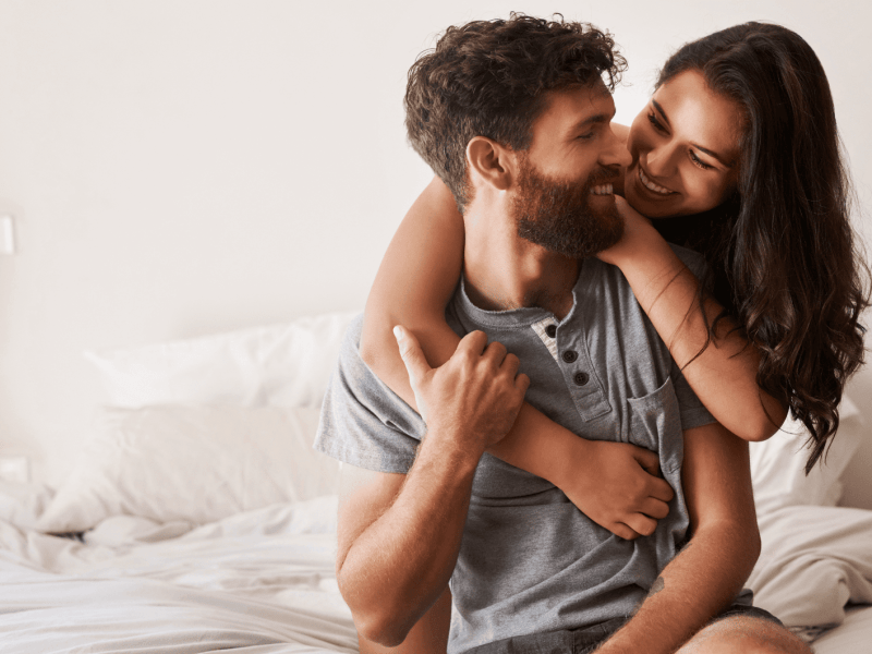 Frau umarmt Mann im Bett von hinten, beide lächeln sich an