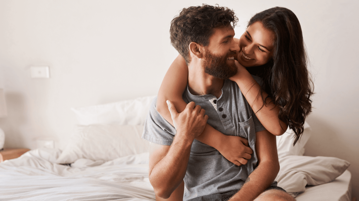 Frau umarmt Mann im Bett von hinten, beide lächeln sich an