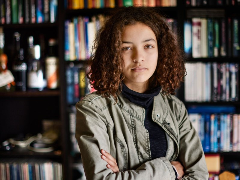 Porträt eines Mädchens, ca. 14 Jahre, das mit verschränkten Armen und ernstem Blick vor einem Bücherregal steht.