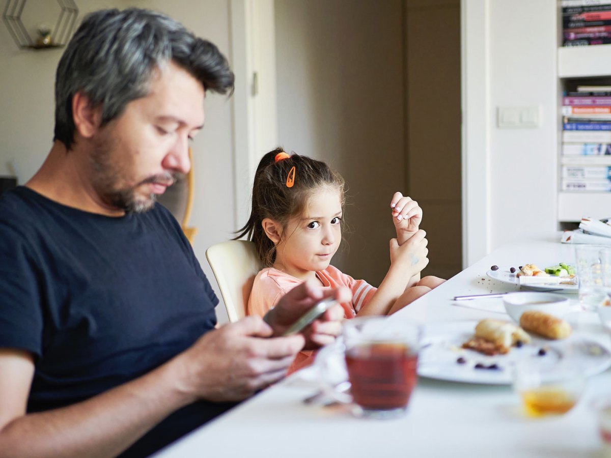 Vater und Tochter sitzen am Fr&uuml;hst&uuml;ckstisch. Der Vater schaut w&auml;hrenddessen auf sein Smartphone.