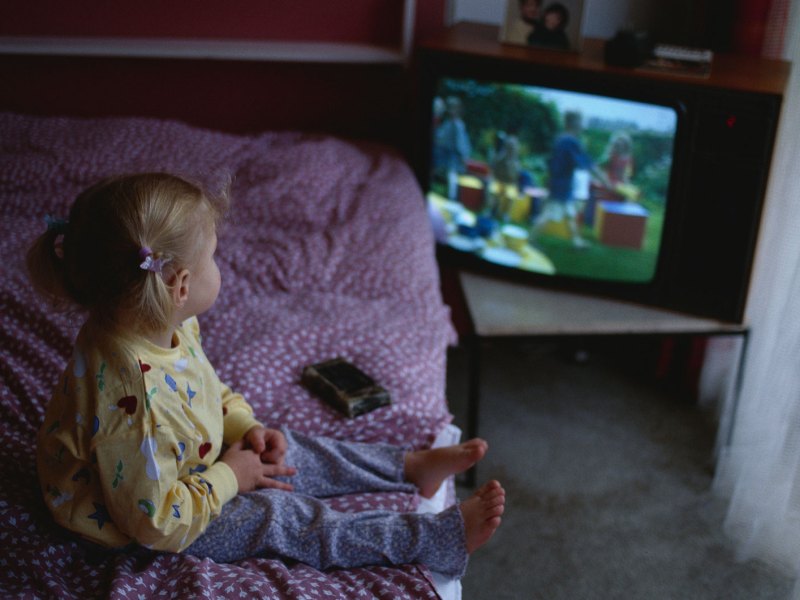 Kleines M&auml;dchen sitzt auf dem Elternbett und schaut etwas auf einem alten R&ouml;hrenfernseher.