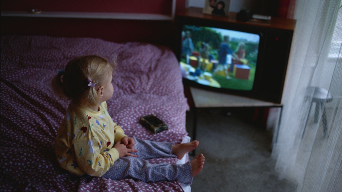 Kleines Mädchen sitzt auf dem Elternbett und schaut etwas auf einem alten Röhrenfernseher.