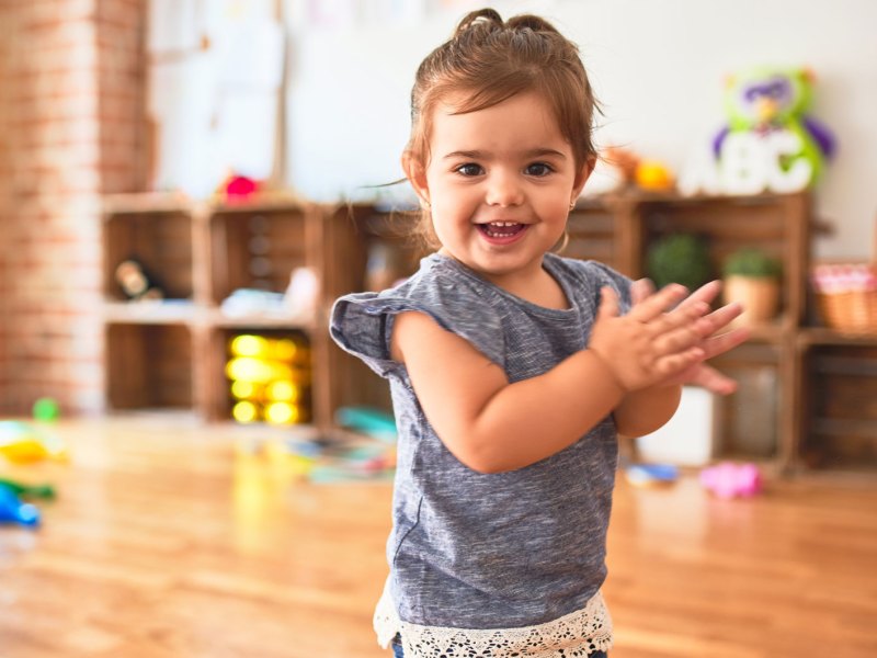 Süßes kleines Mädchen steht in einem großen Raum voller Spielsachen, lacht in die Kamera und klatscht.