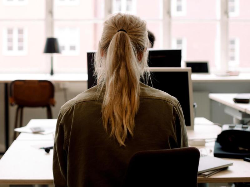 Junge Frau mit einem blonden Pferdeschwanz sitzt an einem Schreibtisch alleine im B&uuml;ro.