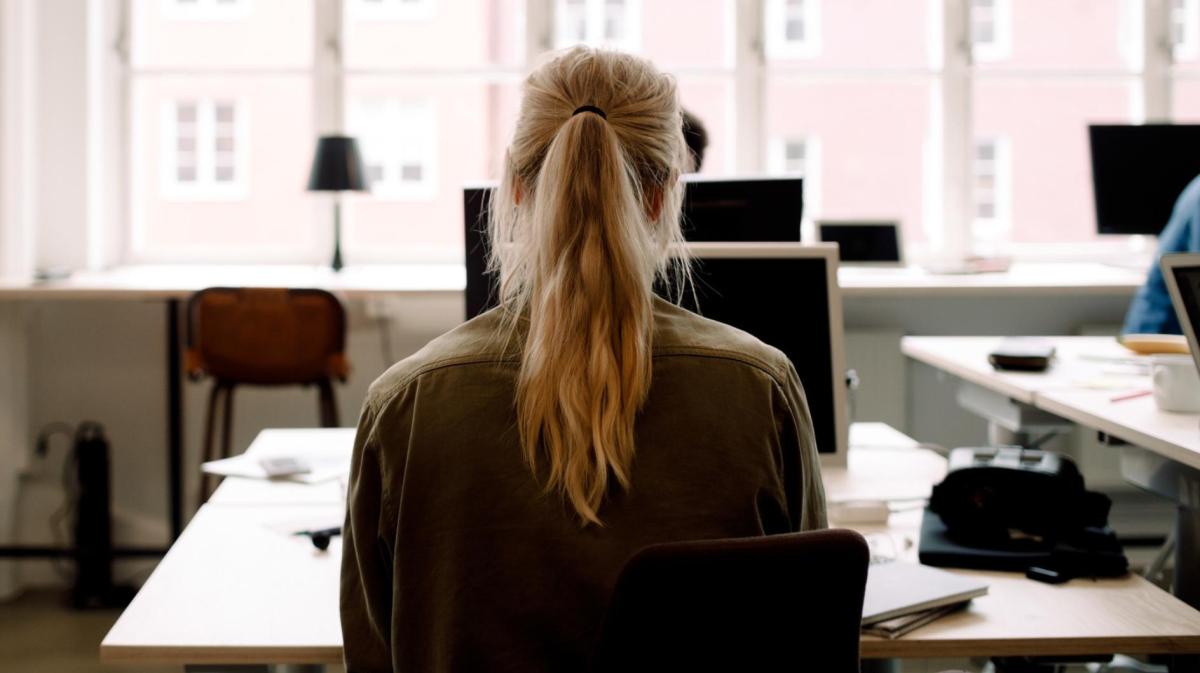 Junge Frau mit einem blonden Pferdeschwanz sitzt an einem Schreibtisch alleine im Büro.