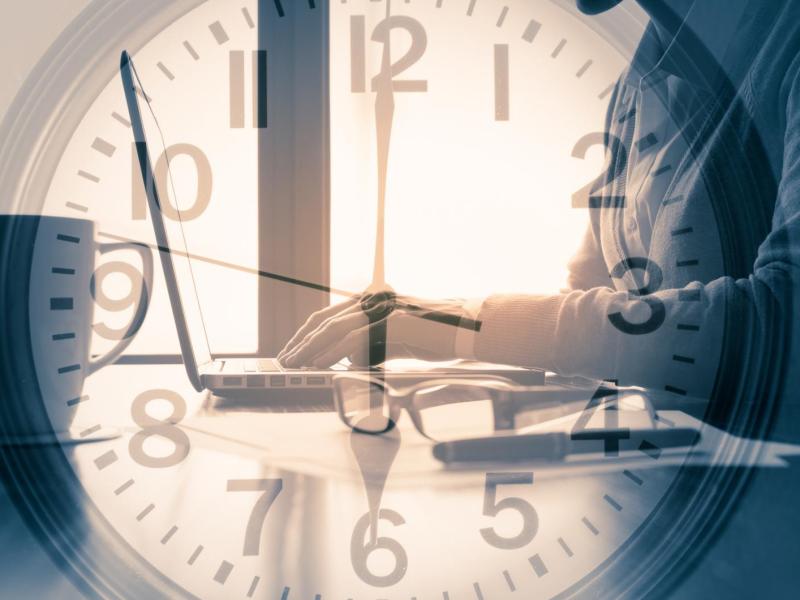 Schattenhafte Frau tippt konzentriert auf Laptop, während große transparente Uhr über dem Bild die Zeit symbolisiert.