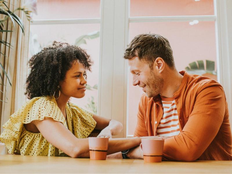 Junge Frau mit Locken in gelber Rüschenbluse hält lächelnd die Hand eines Mannes in orangener Jacke bei einem Kaffee am Fenster.
