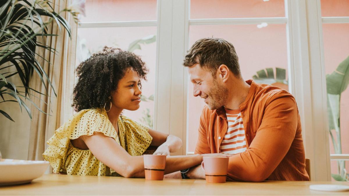 Junge Frau mit Locken in gelber Rüschenbluse hält lächelnd die Hand eines Mannes in orangener Jacke bei einem Kaffee am Fenster.