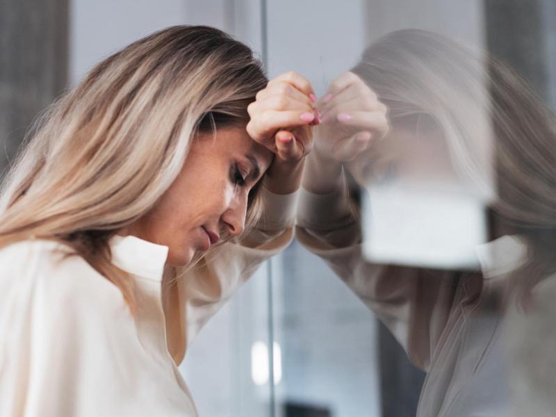 Blonde Frau lehnt sich auf der Arbeit gegen eine Glaswand und stützt dabei ihren Kopf auf ihrem Arm ab.