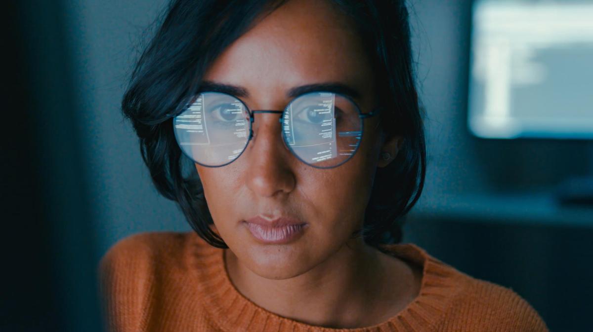 Frau mit dunklen Haaren und Brille sitzt nah vor einem Bildschirm und arbeitet lange.