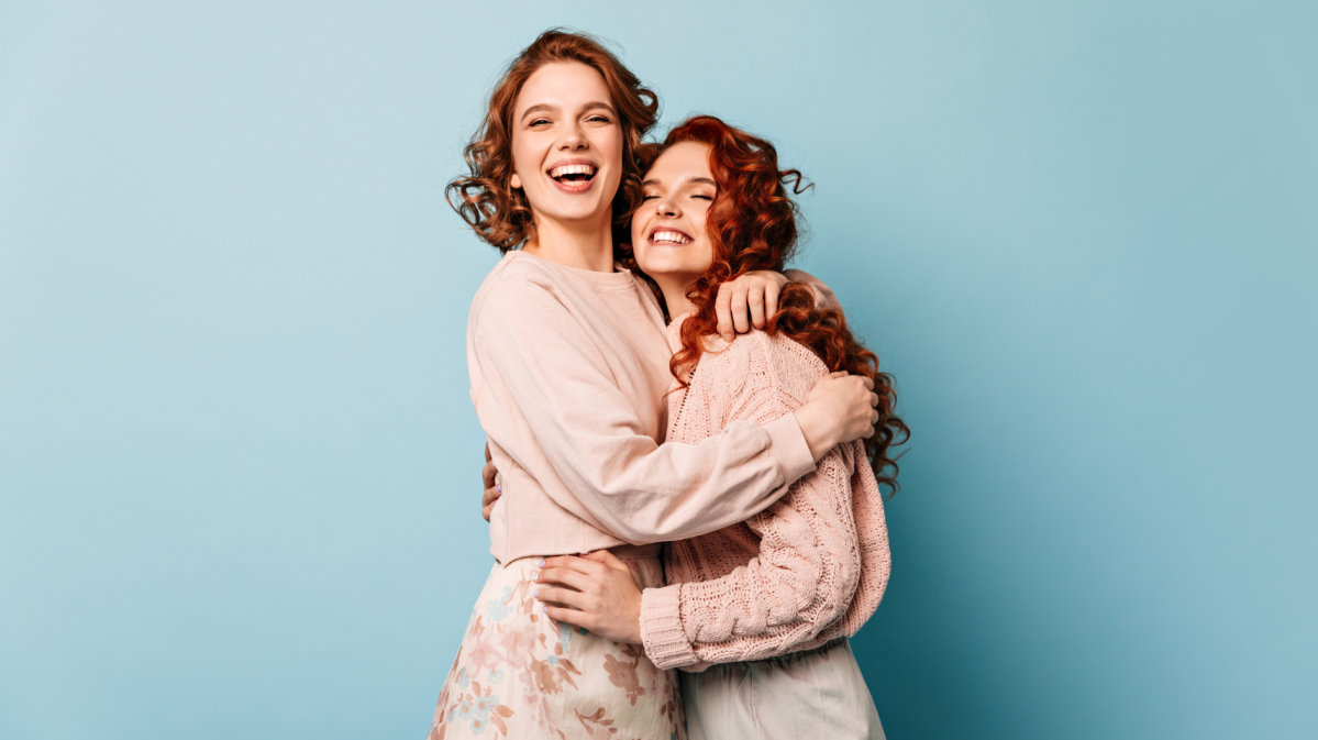 2 junge Frauen stehen vor einem hellblauen Hintergrund, freuen sich und nehmen sich in den Arm