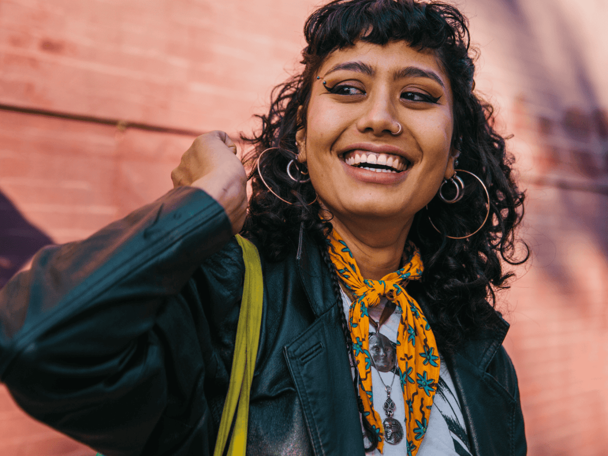 Frau mit dunklen Locken, Piercings und einem Halstuch steht vor einer pinken Wand und strahlt