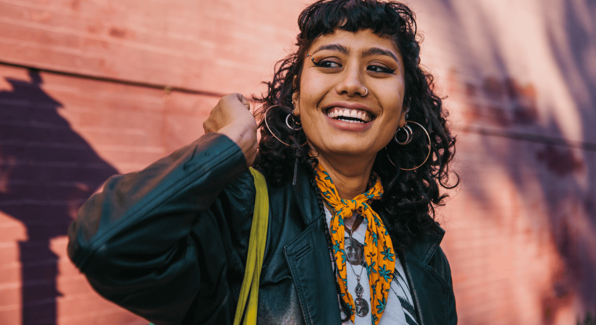 Frau mit dunklen Locken, Piercings und einem Halstuch steht vor einer pinken Wand und strahlt