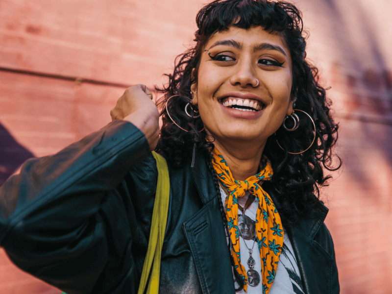 Frau mit dunklen Locken, Piercings und einem Halstuch steht vor einer pinken Wand und strahlt