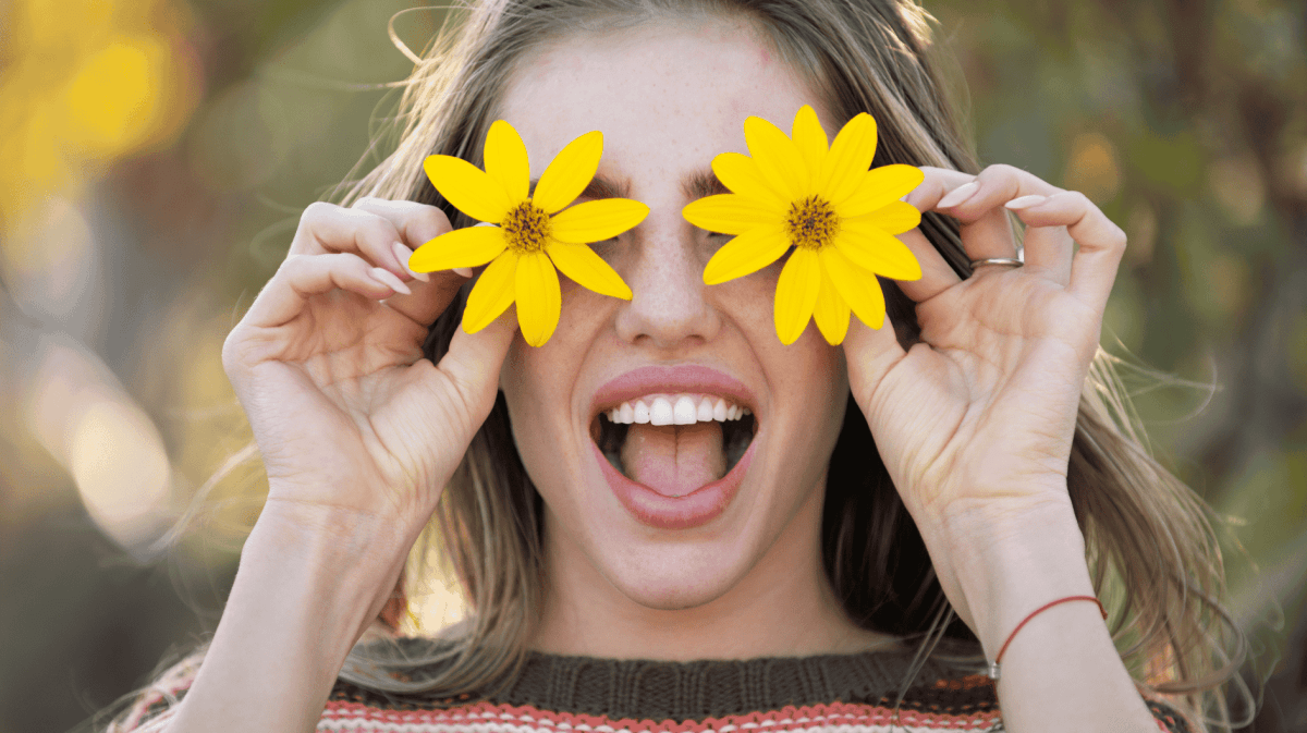 Frau lächelt mit offenem Mund und hält sich zwei gelbe Blüten vor die Augen