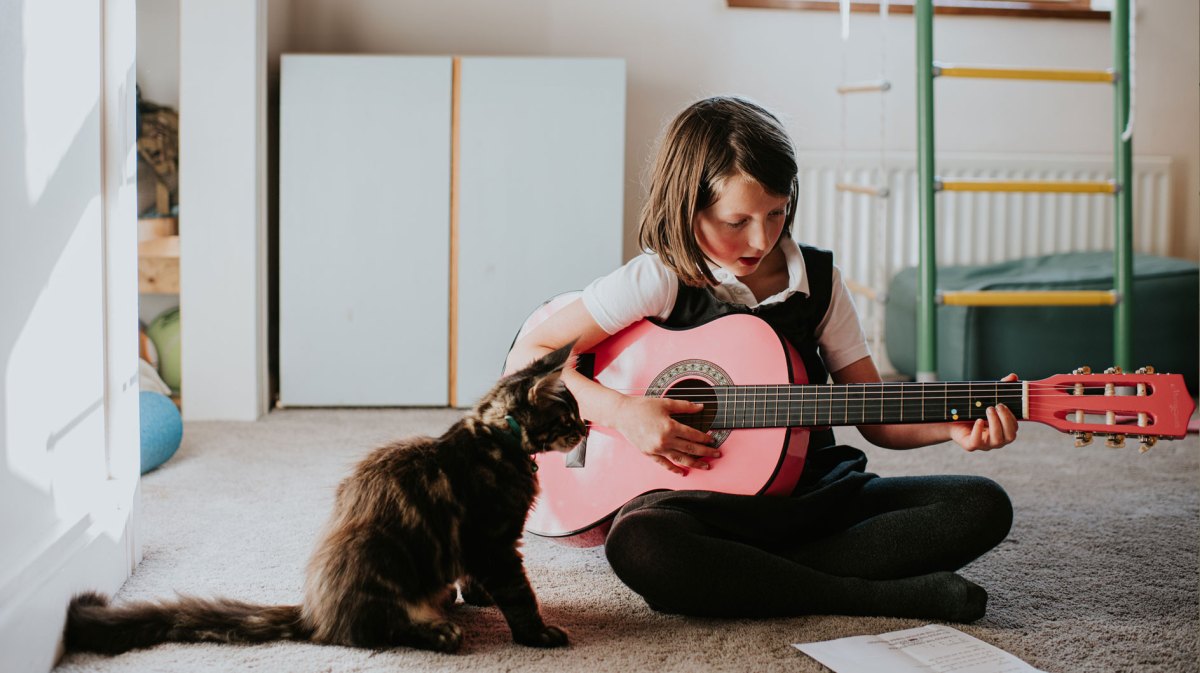 Mädchen sitzt auf dem Fußboden ihres Kinderzimmers und spielt auf einer rosafarbenen Gitarre. Vor ihr sitzt eine kleine Katze.