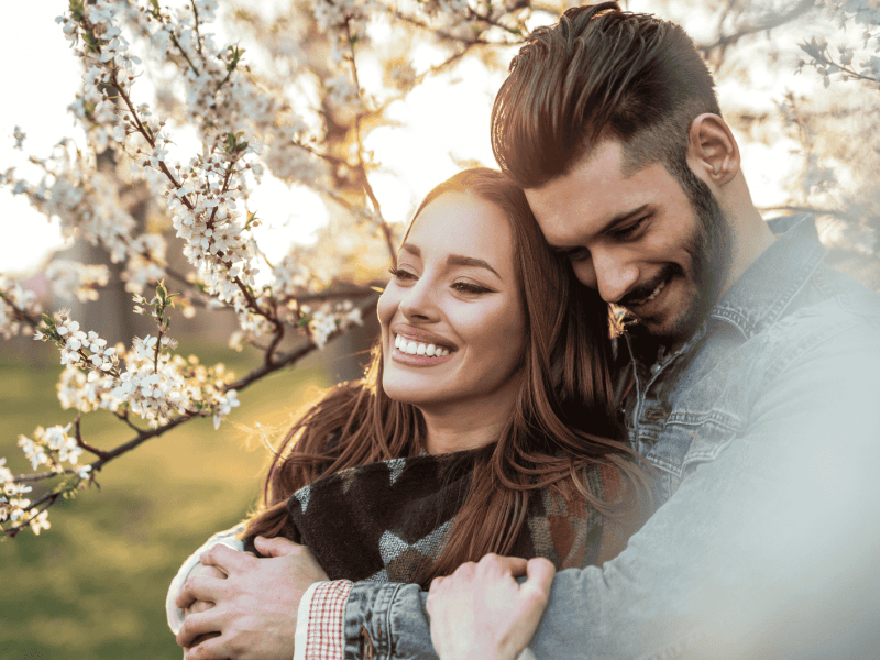 Mann umarmt Frau von hinten, beide lächeln umgeben von Kirschblütenbäumen
