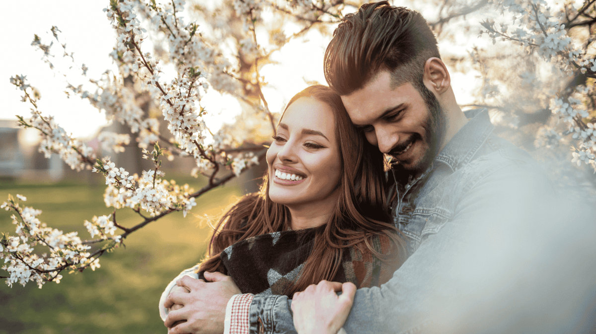 Mann umarmt Frau von hinten, beide lächeln umgeben von Kirschblütenbäumen