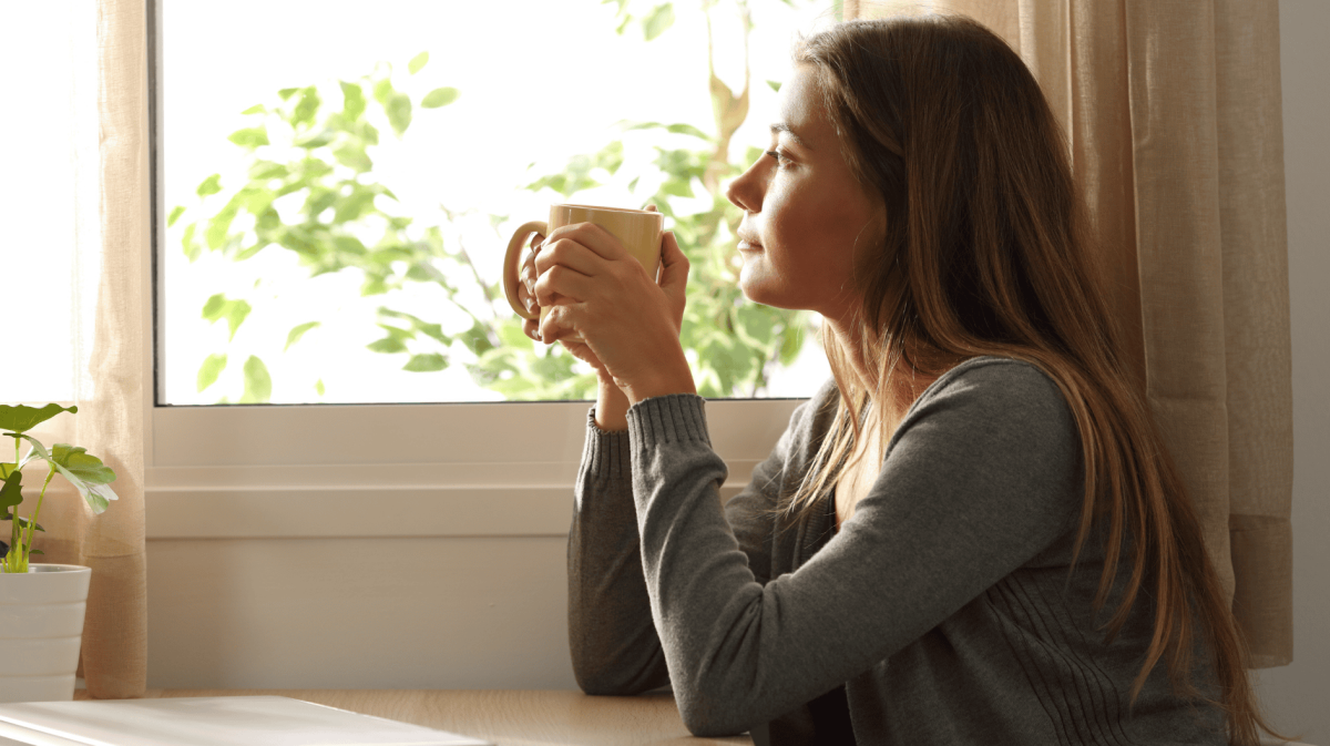 Frau mit Tasse in der Hand schaut nachdenklich aus dem Fenster.