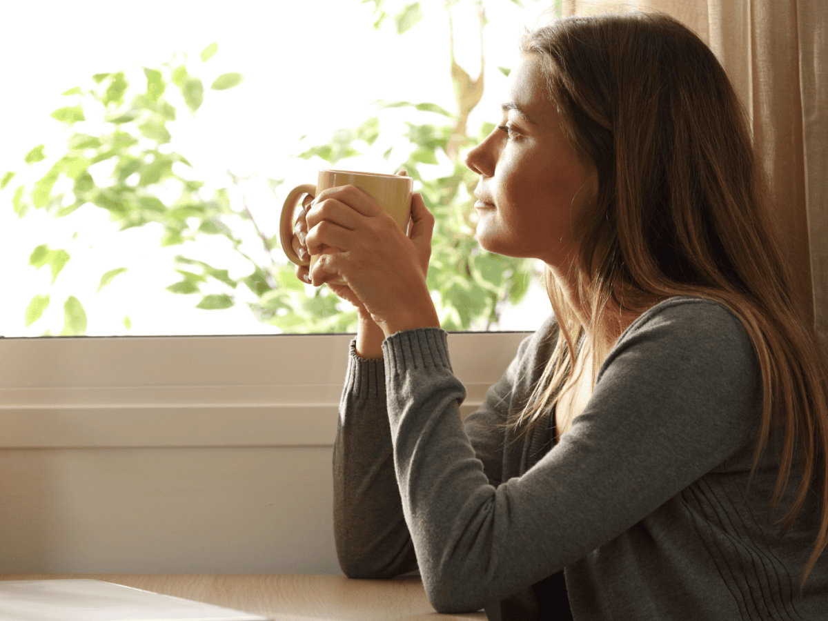 Frau mit Tasse in der Hand schaut nachdenklich aus dem Fenster.