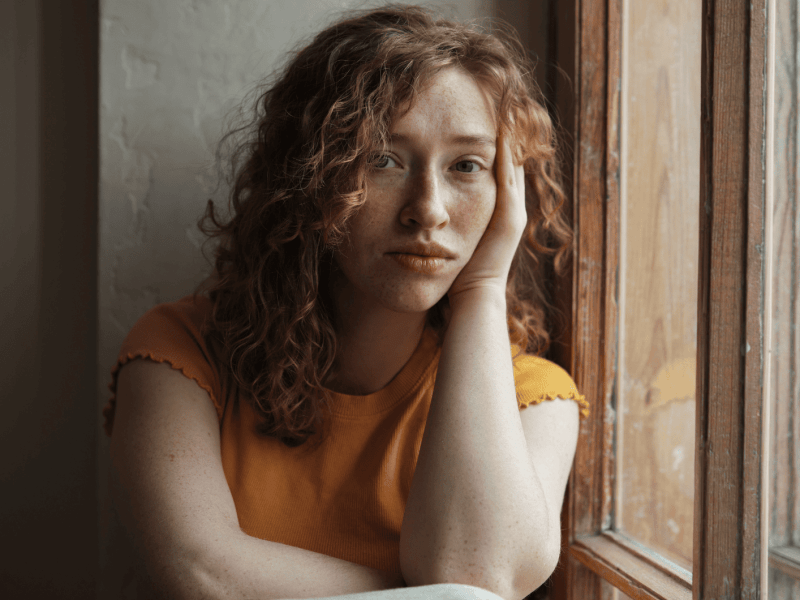Frau mit roten Locken und gelbem T-Shirt sitzt am Fenster und schaut traurig in die Kamera