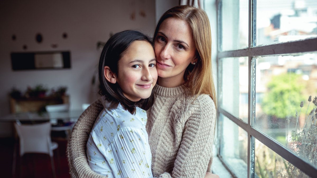 Mutter und Teenagertochter umarmen sich vor einem Fenster. Die Tochter lacht entspannt, die Mutter sieht besorgt aus.