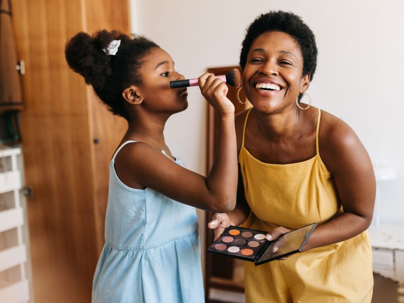 Tochter, ca. 10 Jahre alt, trägt Rouge auf die Wange der Mutter auf. Beide stehen im Schlafzimmer.