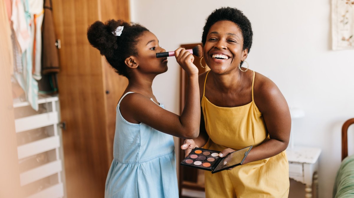 Tochter, ca. 10 Jahre alt, trägt Rouge auf die Wange der Mutter auf. Beide stehen im Schlafzimmer.