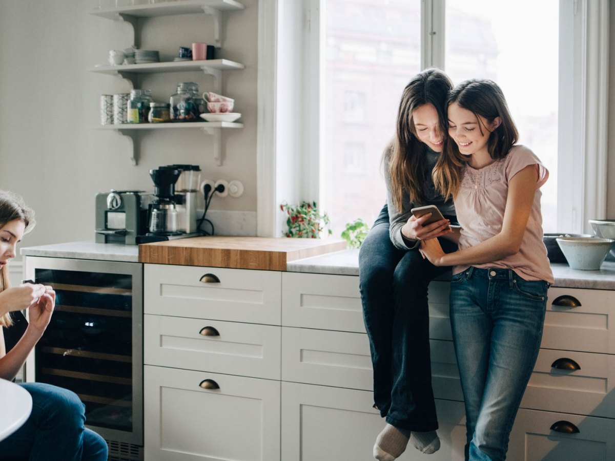 Drei Teenager-M&auml;dchen in der K&uuml;che, schauen etwas auf ihren Smartphones nach.