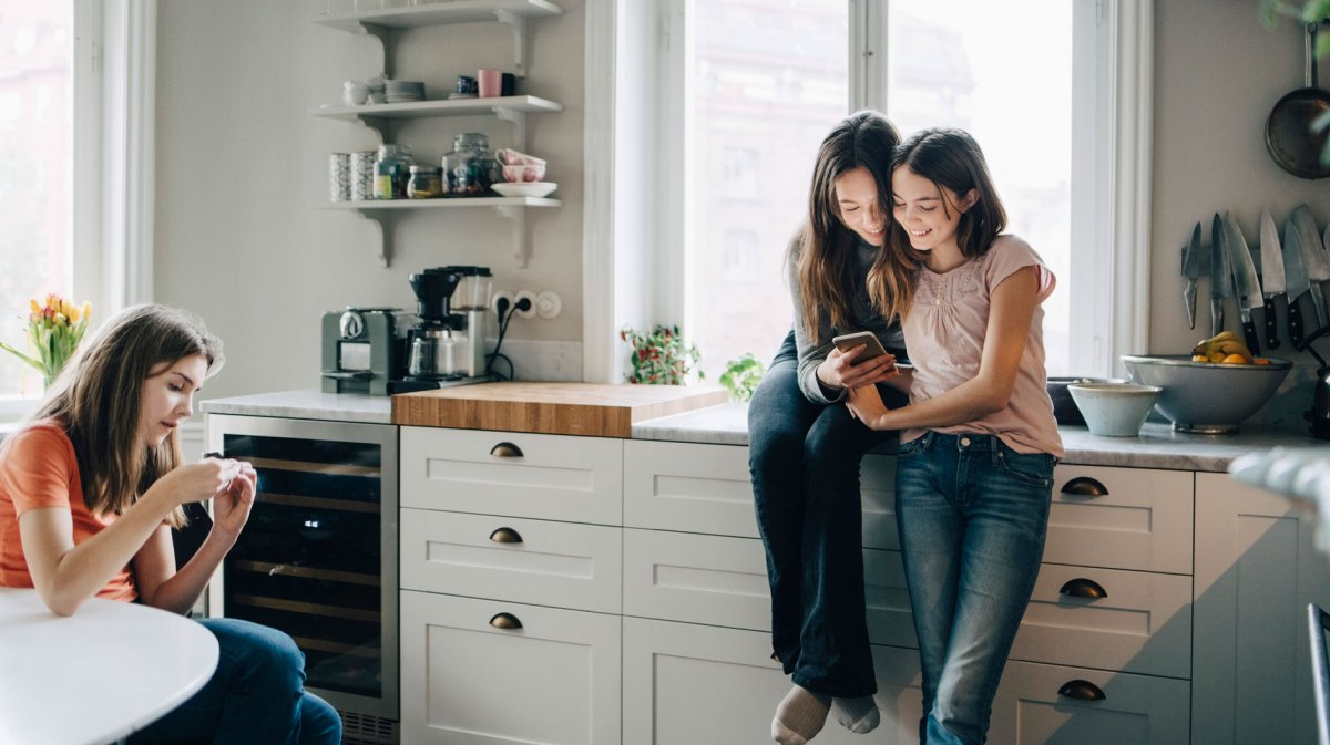 Drei Teenager-Mädchen in der Küche, schauen etwas auf ihren Smartphones nach.