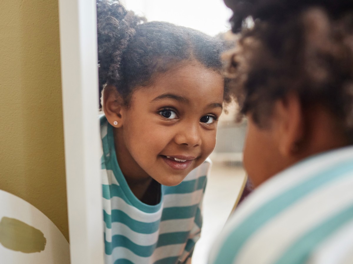 Nahaufnahme eines Mädchens, das spielerisch über einen Spiegel in die Kamera lacht.