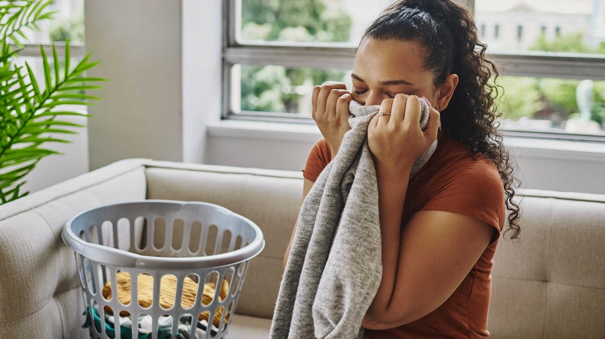Junge Frau mit lockigem, dunklem Haar riecht lächelnd an grauem Pullover, neben ihr ein Wäschekorb auf beigem Sofa.