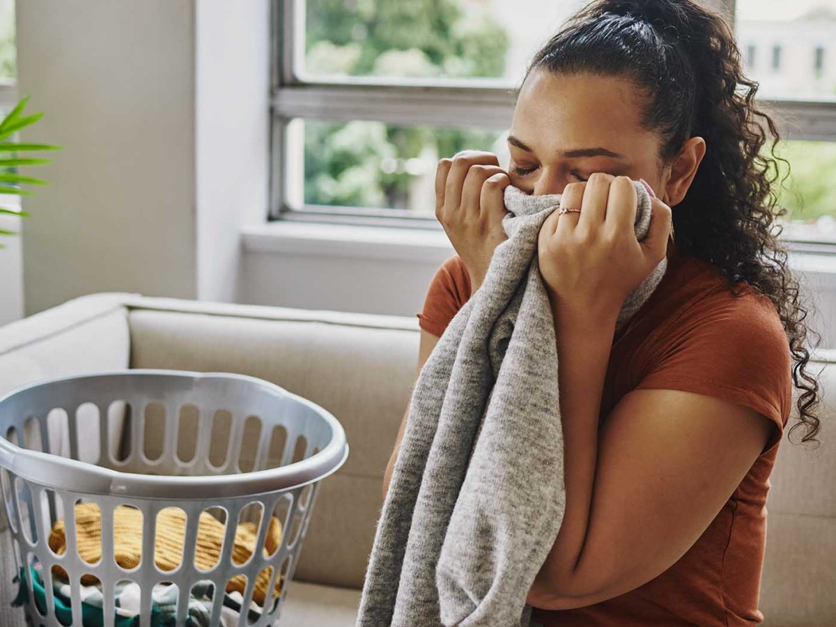 Junge Frau mit lockigem, dunklem Haar riecht lächelnd an grauem Pullover, neben ihr ein Wäschekorb auf beigem Sofa.