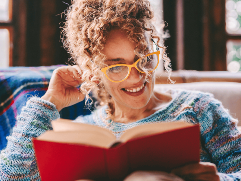 Frau mit Locken sitzt auf Sofa und liest lächelnd ein Buch.