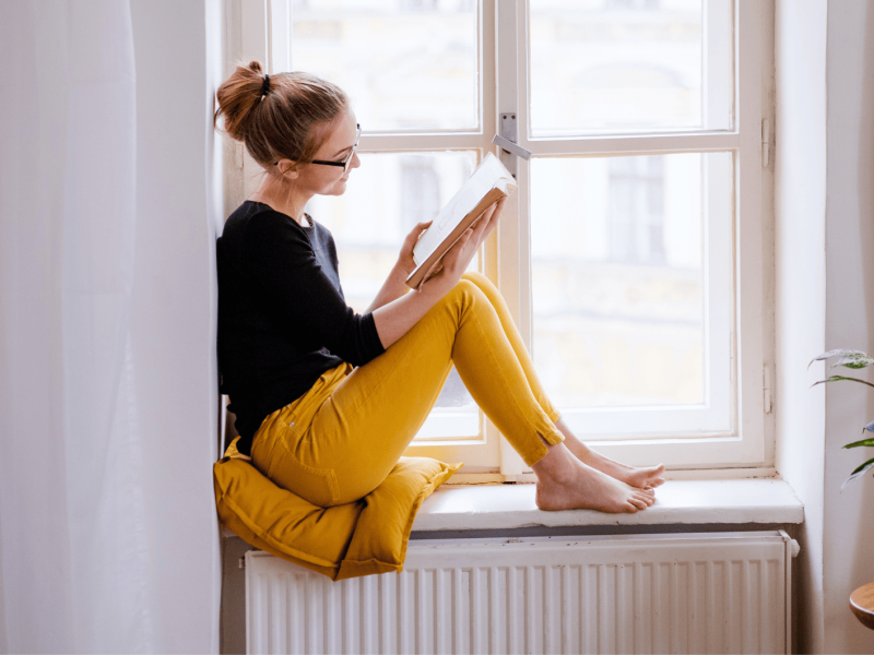 Frau sitzt auf Fensterbank und liest ein Buch.
