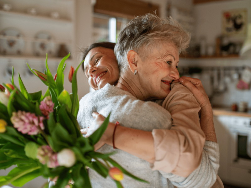 Junge Frau umarmt ihre Mutter und &uuml;berreicht ihr Blumenstrau&szlig;.