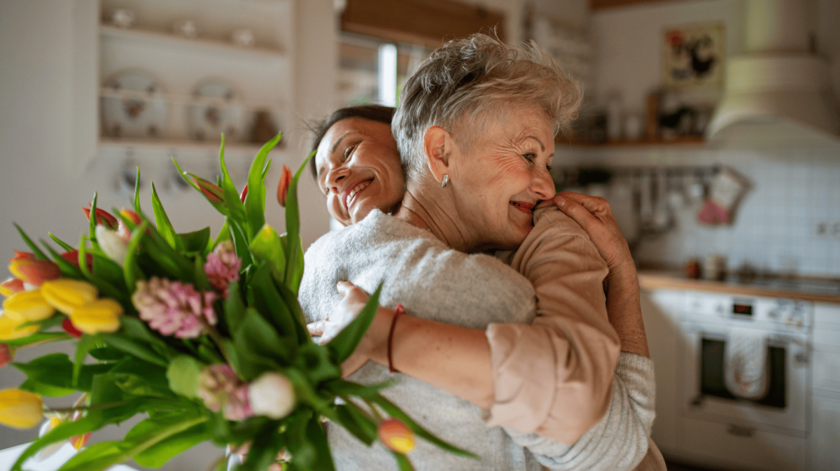 Junge Frau umarmt ihre Mutter und überreicht ihr Blumenstrauß.