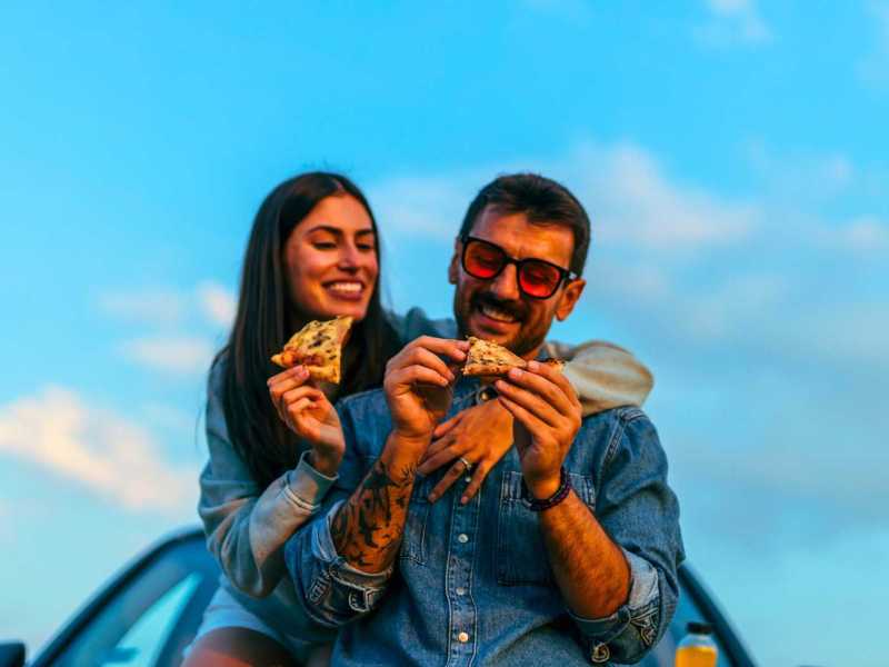 Paar sitzt auf einem Autodach und isst Pizza. Hinter ihnen strahlend blauer Himmel.