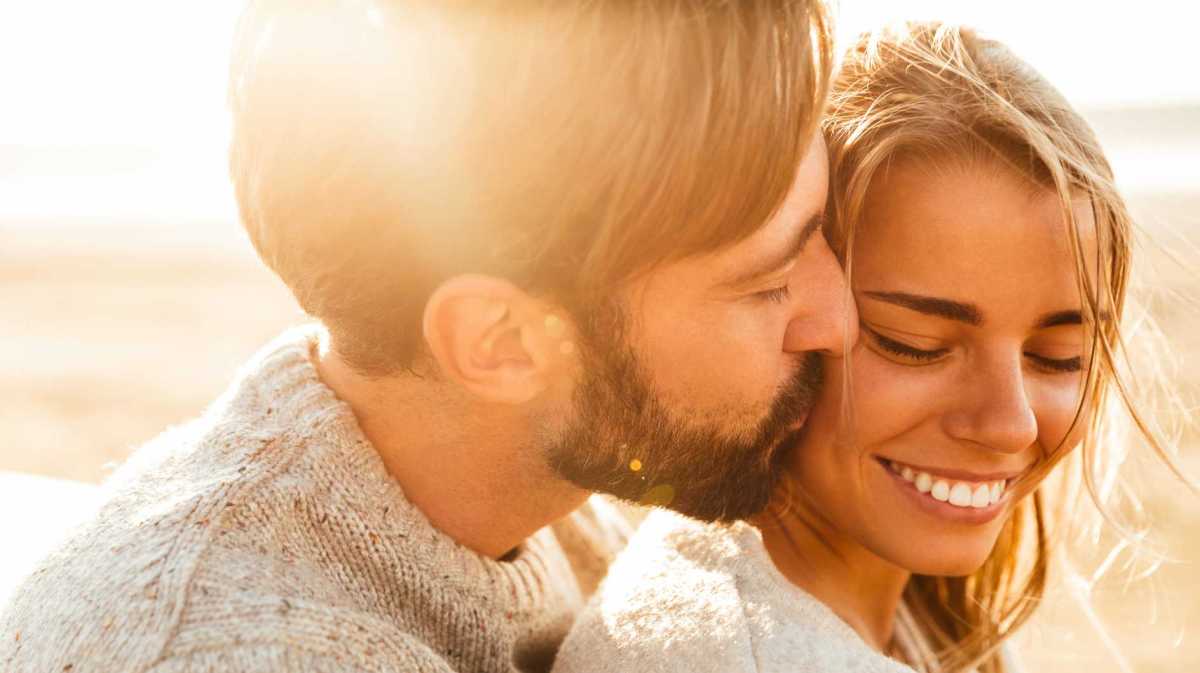 Mann umarmt seine Freundin und gibt ihr einen Kuss auf die Wange. Sie lächelt.
