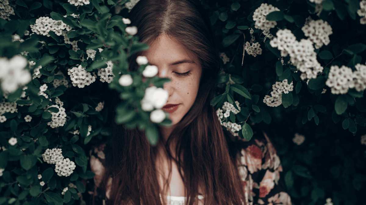 Brünette Frau steht vor Gebüsch mit Blumen.
