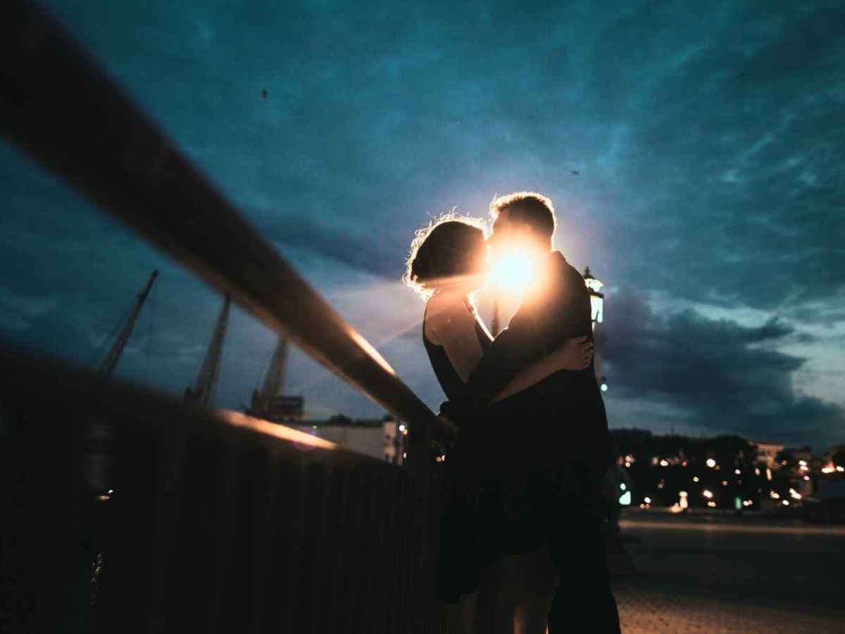 Paar steht nachts auf einer Br&uuml;cke, hinter ihnen leuchtet ein helles Licht, sie sind innig und k&uuml;ssen sich.