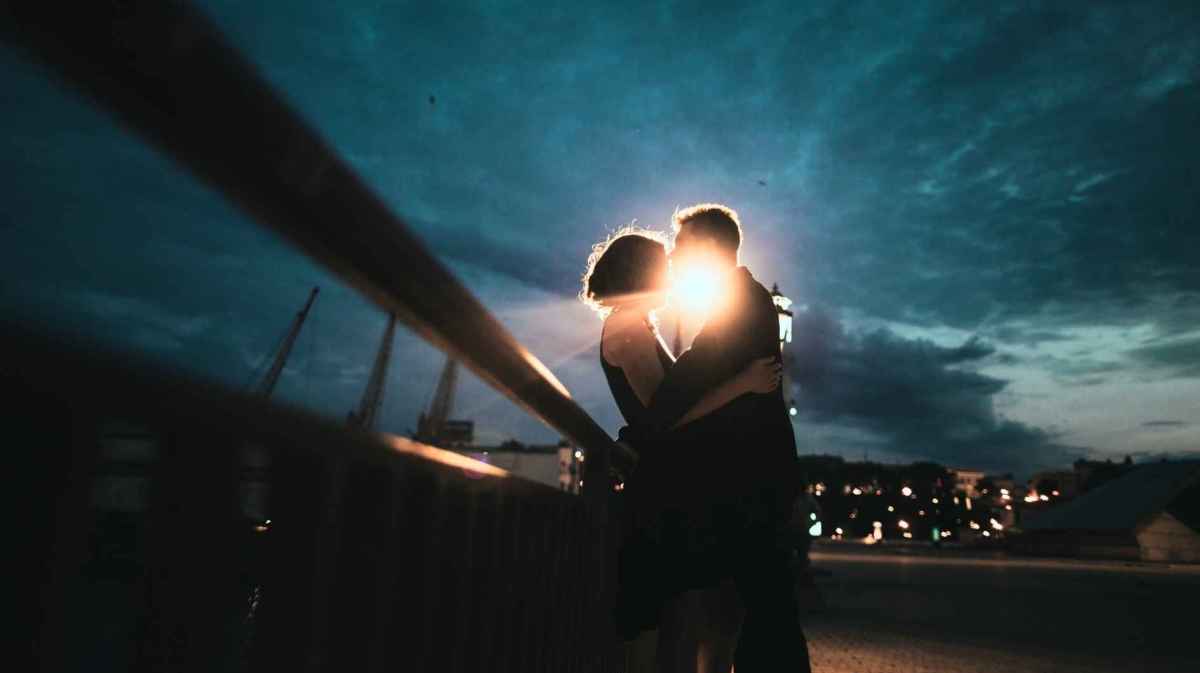 Paar steht nachts auf einer Brücke, hinter ihnen leuchtet ein helles Licht, sie sind innig und küssen sich.