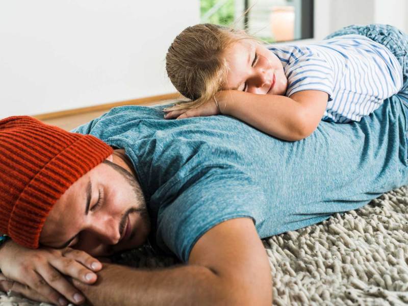 Vater und Tochter liegen bäuchlings auf dem Teppich, die Tochter auf dem Rücken des Vaters.