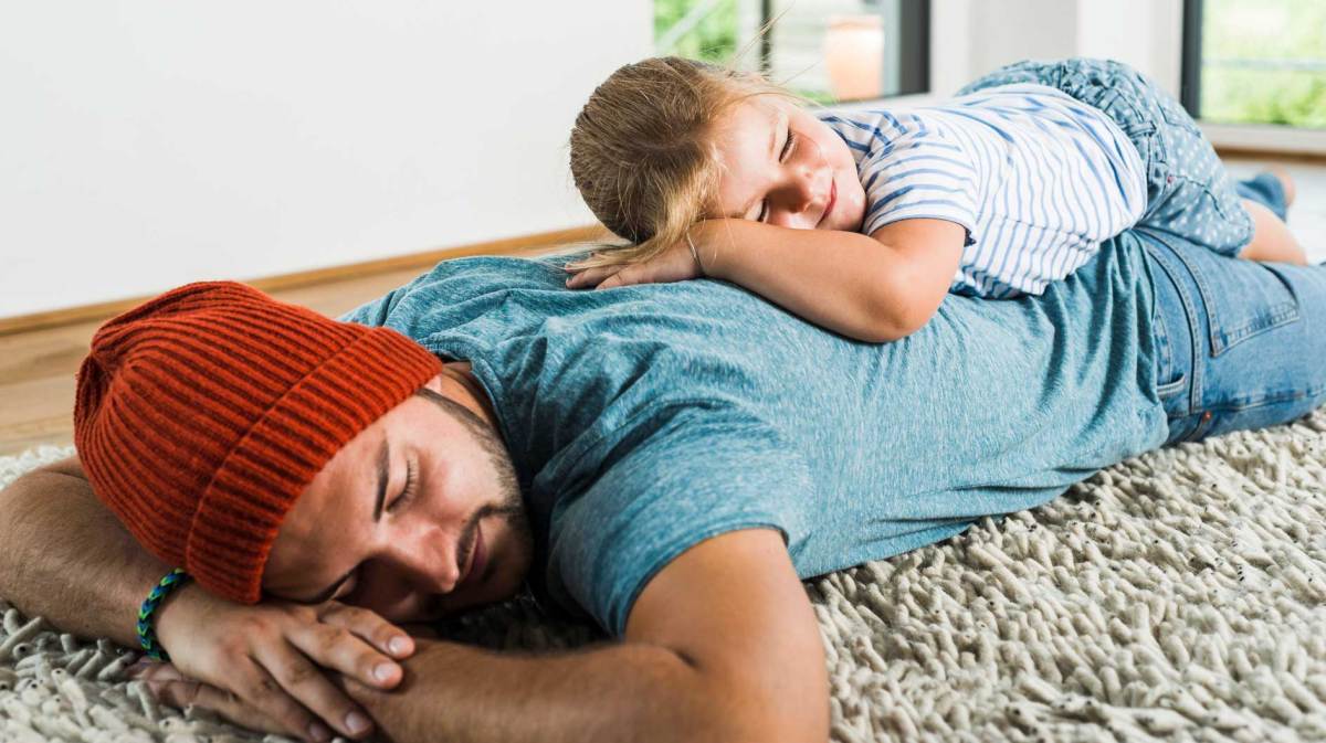 Vater und Tochter liegen bäuchlings auf dem Teppich, die Tochter auf dem Rücken des Vaters.